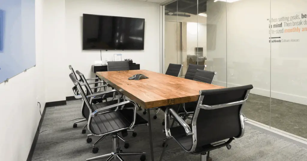 Private meeting room at Strive Coworking Workspaces with wood conference table, ergonomic chairs, glass walls, and wall-mounted display screen.